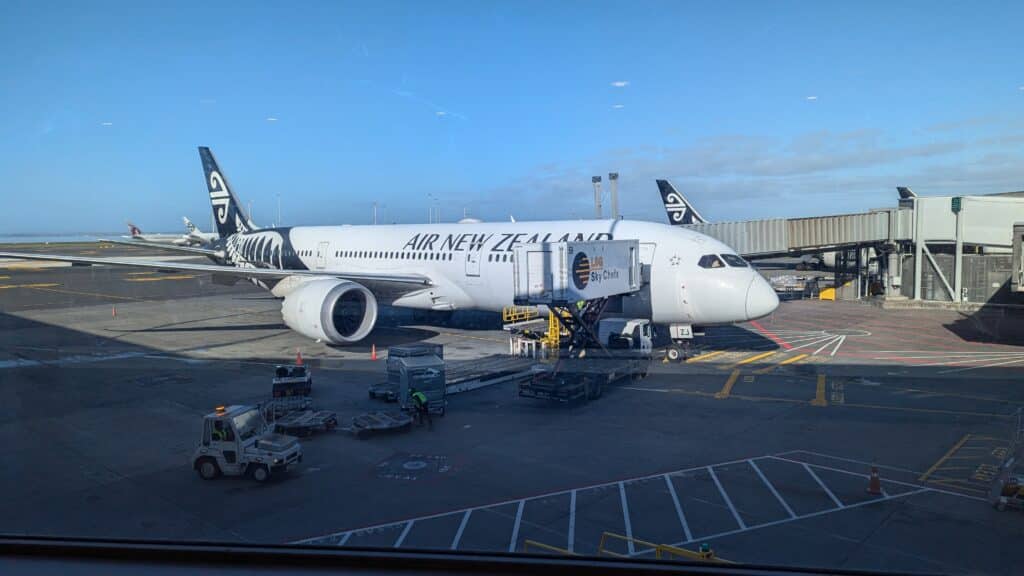 紐西蘭航空飛機在停機坪與機場航廈連接的照片，行李正在搬運、旅客行走廊道通行中 。